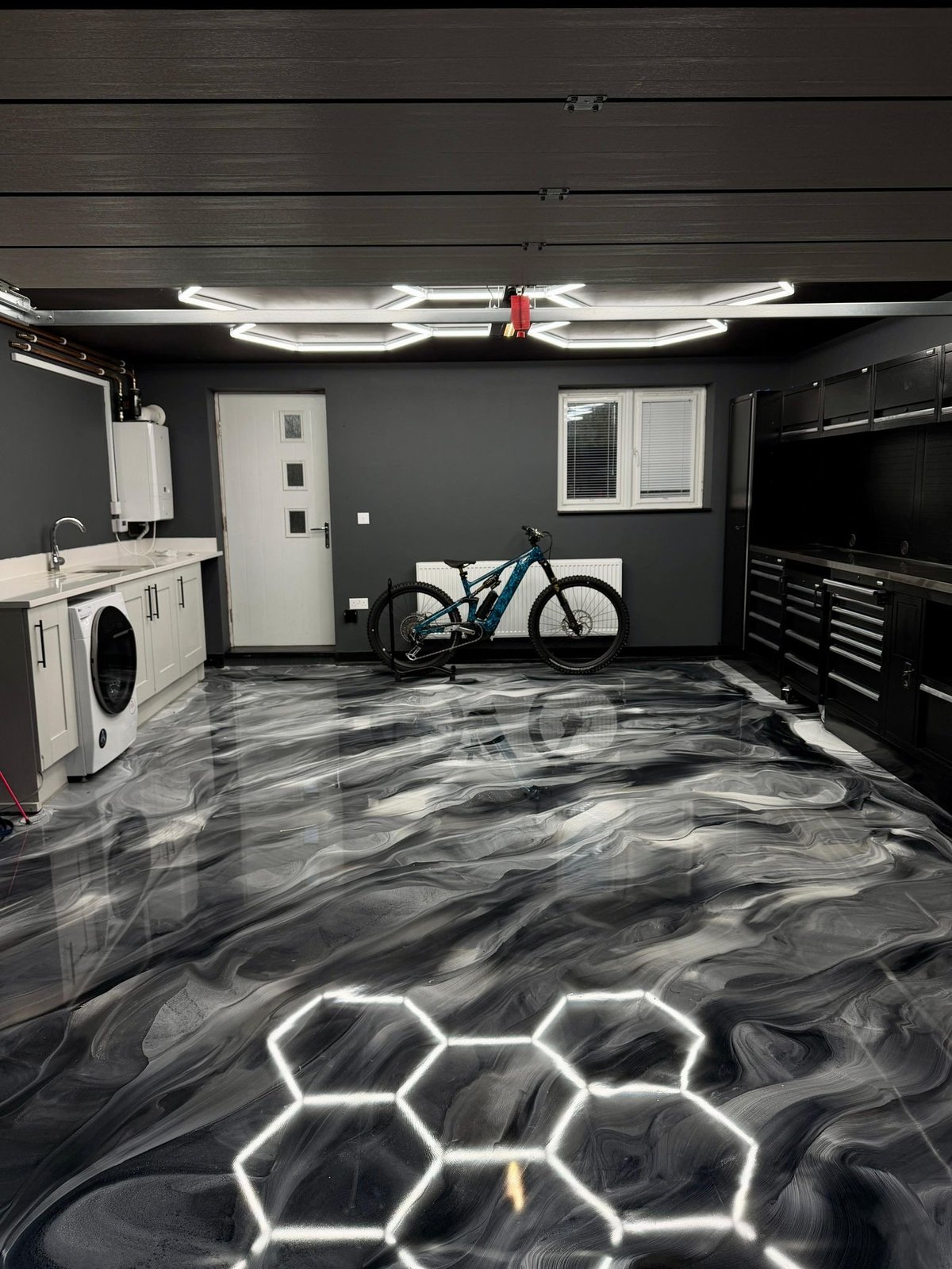 Modern garage interior with black marble flooring, neon lighting, blue mountain bike, white washer/dryer, tool cabinet, and bright ceiling panels